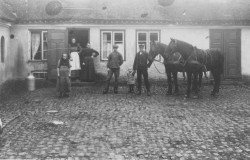 Kvinderne ved køkkendøren, mændene ved hestene. Tjenestepigen til venstre i billedet står isoleret for sig selv.
Fotografiet er brugt som en julehilsen og poststemplet i 1909.